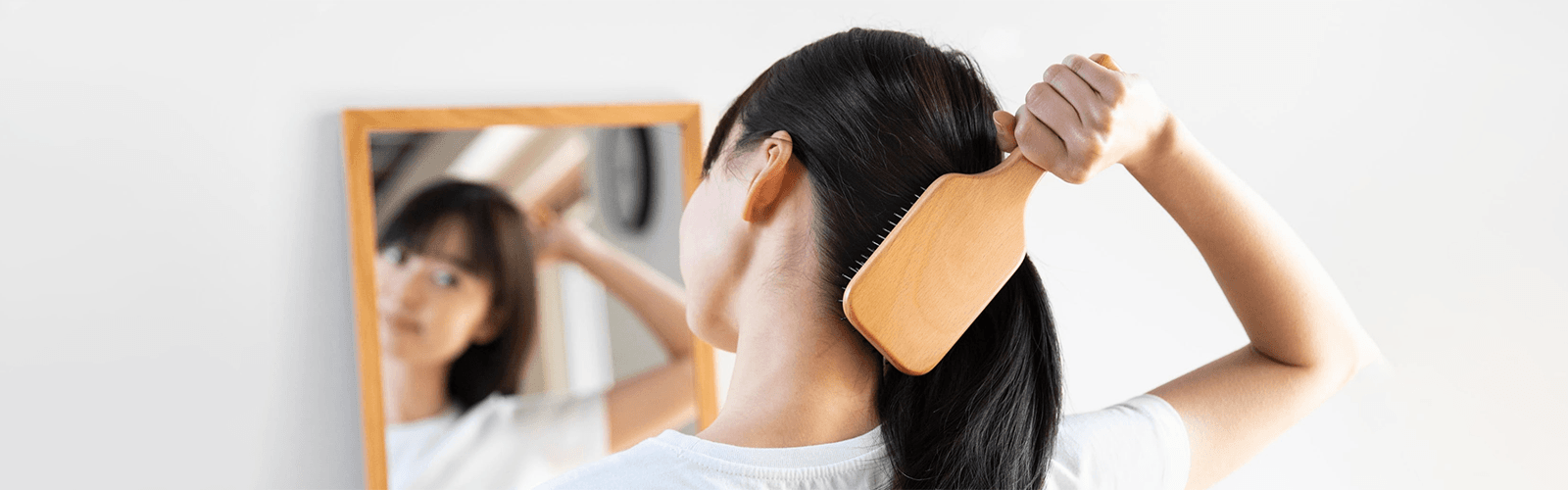 Woman with long black hair standing in front of a mirror brushing her hair with a MUJI Brush. 