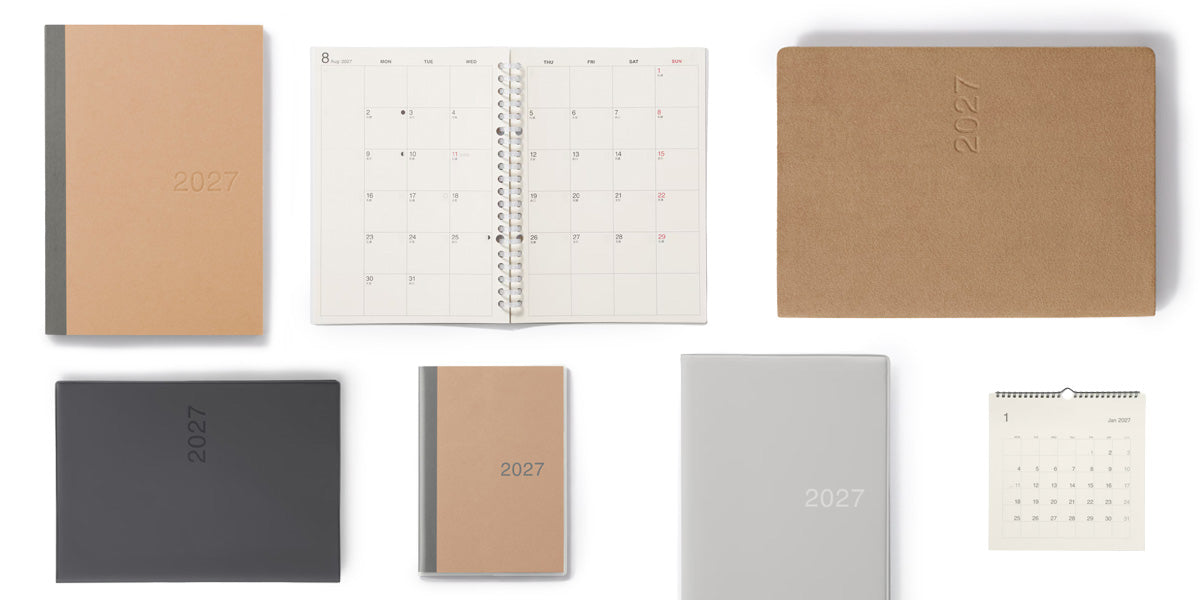 A set of minimalist 2027 planner and calendar products arranged on a white background, including closed notebooks in tan, dark gray, and light gray, an open monthly calendar, a large tan cover, a small wall calendar, and a tan 2027 journal.