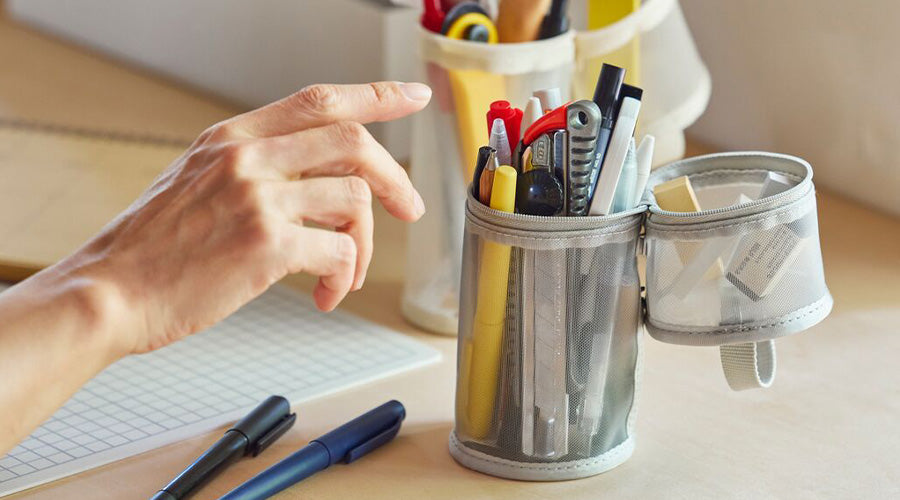 Hand reaching for a pen in a pen case on a desk with stationery items.