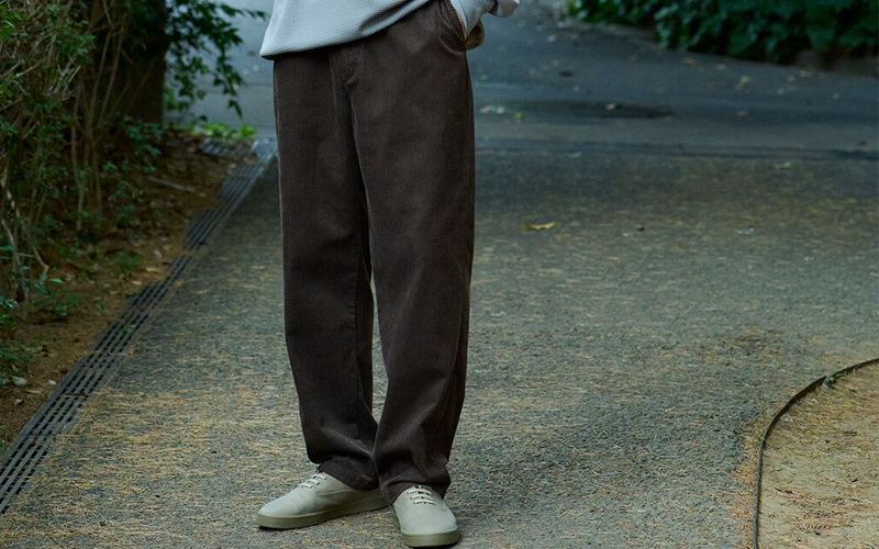 Person wearing brown pants and light-colored shoes on a sidewalk with greenery.