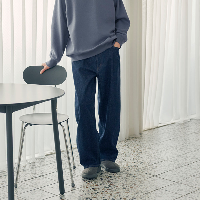 A person wearing a loose gray sweatshirt and dark blue straight-leg jeans stands beside a black table in a bright room with white curtains and tiled flooring.