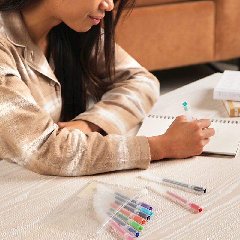 Person writing in a notebook with colorful pens on a wooden table.