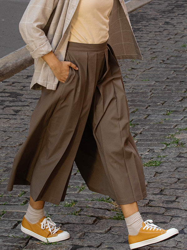 Person wearing a beige sweater, brown wide-leg pants, and yellow less tiring sneakers on a cobblestone street.