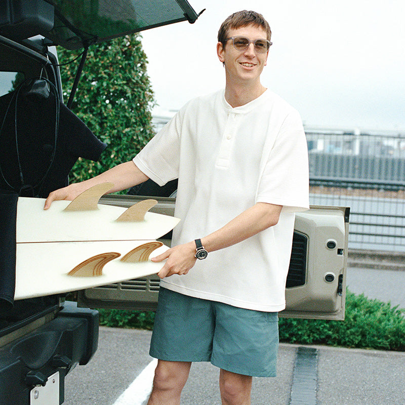 Man wearing a MUJI t-shirt & shorts grabbing a surf board out of the trunk of a car. 