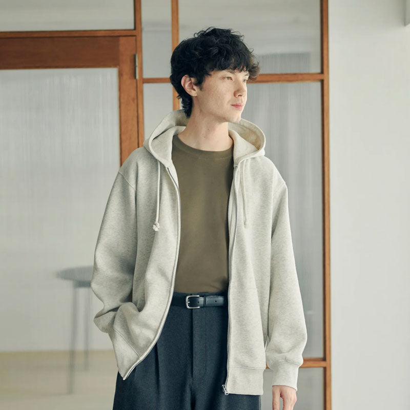 A man wearing a light gray zip-up hoodie over a brown t-shirt and dark gray trousers