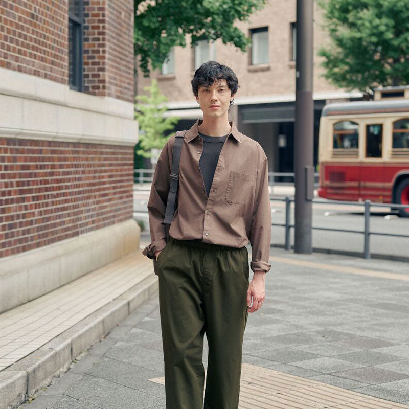 A man stands on a city sidewalk wearing a brown button-up shirt